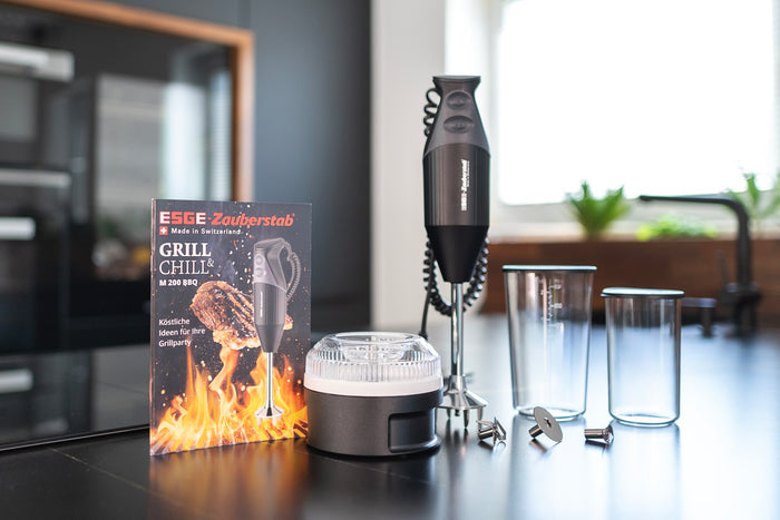 Der Unold ESGE-Zauberstab® M 200 BBQ Stabmixer mit Aufsätzen, zwei durchsichtigen Behältern und einem Rezeptheft mit der Aufschrift GRILL & CHILL steht auf einer Küchentheke, im Hintergrund sind ein unscharfes Fenster und eine Spüle zu sehen.
