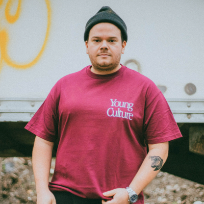 A man in a maroon Young Culture T-shirt and black beanie stands outdoors, showing a tattoo and watch. The blurred background with graffiti hints makes this look perfect for fall street style.