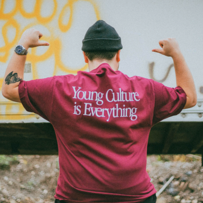 A person wearing a maroon Young Culture t-shirt with “Young Culture is Everything” on the back flexes for the camera, paired with a black beanie and showing off a left arm tattoo—an ideal look for fall.