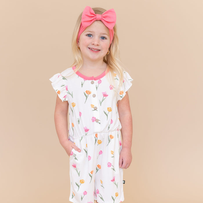Young girl posing wearing the Flutter Sleeve Romper in Tulip paired with a pink bow