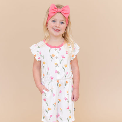 Young girl posing wearing the Flutter Sleeve Romper in Tulip paired with a pink bow