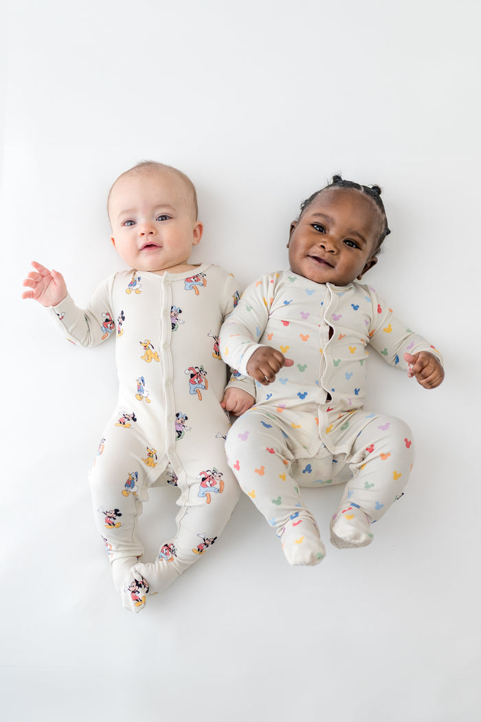 Two babies wearing colorful mickey mouse footed onesies on a white background