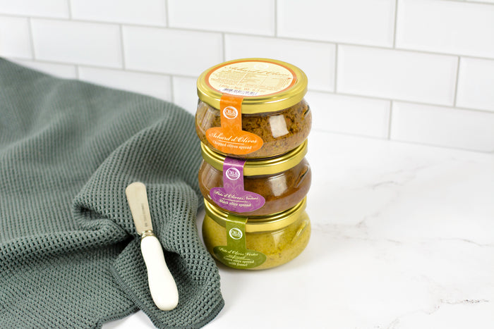 A set of three glass jars containing different flavors of olive tapenade stacked together next to a white ceramic butter knife, with a green cloth napkin and white tiled background.