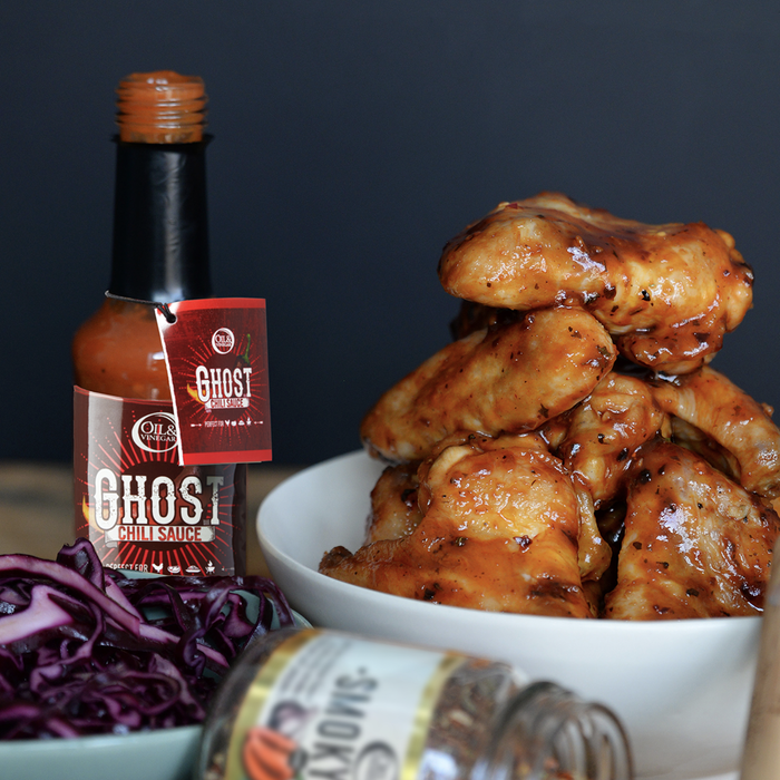 Chicken wings with sauce on them in a white bowl. Bottle of Oil & Vinegar Ghost chili sauce with a red label beside it