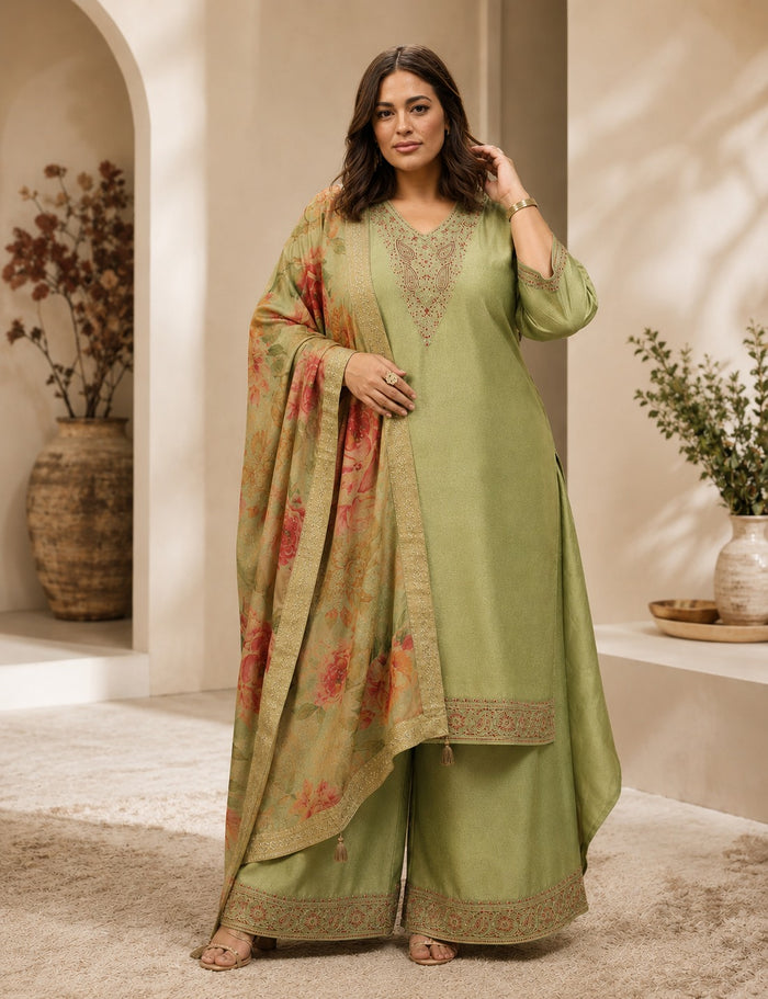 A woman models a sage green three-piece traditional outfit with matching floral dupatta in a warm, sunlit indoor setting with beige walls and arches.