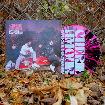 We see a vinyl cover with a picture of the band members sitting on an old 50s Cadillac. It is nighttime in the middle of winter and the stars are visible behind the band. In the top left corner there is text that reads Cherie Amour Internal Discussions. Sticking out of the vinyl cover is a vinyl colored pink and black. The album is leaning against a chain link fence in the middle of fall. There are leaves on the ground.