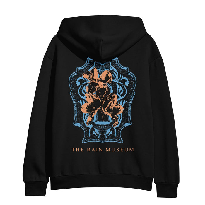 The back of the hoodie is a blue and orange colored crest design with THE RAIN MUSEUM written in orange text below it.