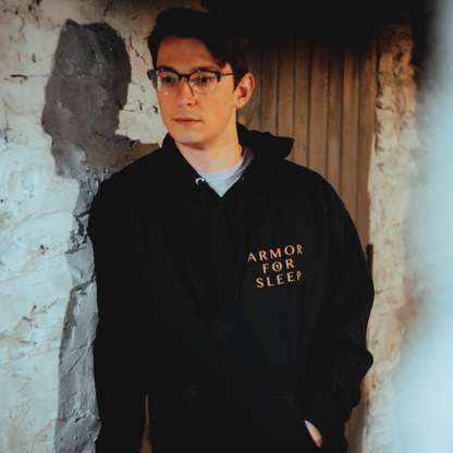 Someone wearing Front of black pullover hoodie with ARMOR FOR SLEEP written in orange text in the top corner of the chest.
