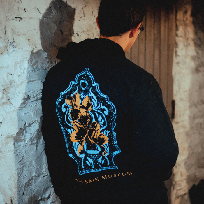 A person with short dark hair and glasses wears an Armor For Sleep pullover hoodie with a blue and gold ornate floral design and "THE RAIN MUSEUM" on the back, standing in front of a weathered wall and wooden door.