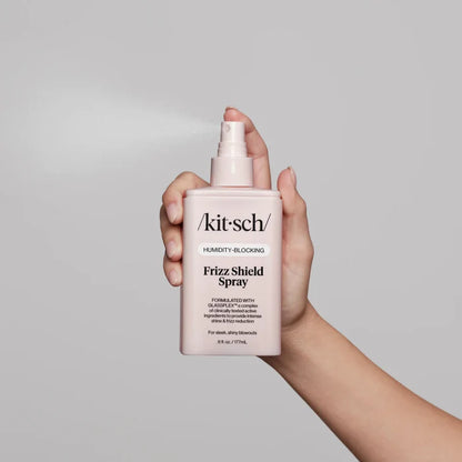 Hand holding a pink bottle of 'kitsch' Frizz Shield Spray against a gray background