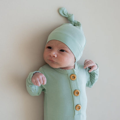Close up of newborn wearing Kyte Baby Knotted Gown with Hat Set in Sage  while laying on a crib sheet in Oat