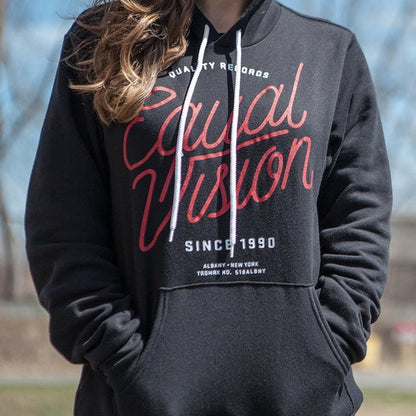 Black Equal Vision hoodie. Center of hoodie has "EQUAL VISION" printed in red cursive. Above has "QUALITY RECORDS" printed in white and below has "SINCE 1990" printed in white. Individual is modeling hoodie outside with blue sky and trees in the background.