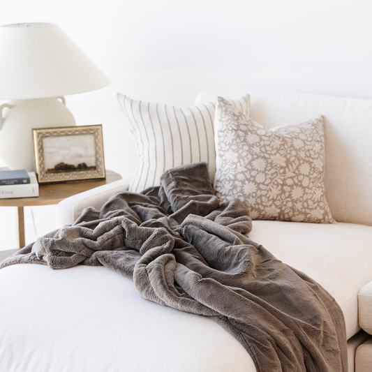 A brown luxury lush Saranoni blanket is draped on a couch. This cozy blanket is a luxury blanket and a faux fur blanket. This soft blanket is great as a home decor staple, comfort gift, or everyday comfort item. Elevate your home and home decor with this faux fur blanket that is also a throw blanket. Enjoy this luxury blanket for years to come!