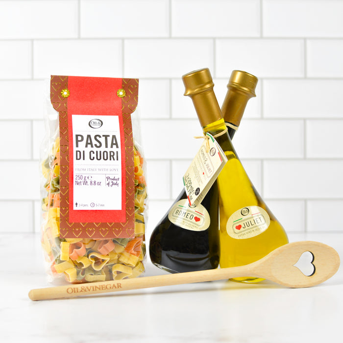 A gift set of culinary items including a package of heart-shaped pasta (Pasta di Cuori), a wooden spoon with a heart-shaped cutout, and bottles of extra virgin olive oil and artisanal balsamic vinegar.