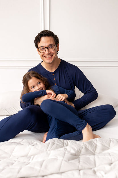 Man and child in matching navy pajamas sitting on a bed.