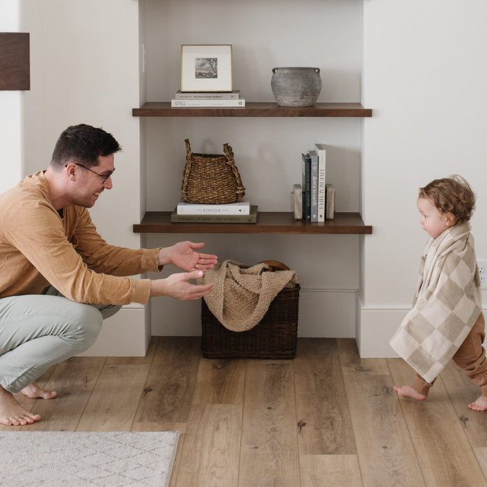 A little boy walks to his dad and has a neutral checkered minky stretch blanket around his shoulders. The soft blanket is a luxury blanket and a Saranoni blanket. This cozy blanket is also a receiving blanket and is a cute and cozy baby blanket. This stretchy blanket is a great nursery item.