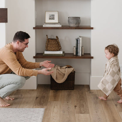 A little boy walks to his dad and has a neutral checkered minky stretch blanket around his shoulders. The soft blanket is a luxury blanket and a Saranoni blanket. This cozy blanket is also a receiving blanket and is a cute and cozy baby blanket. This stretchy blanket is a great nursery item.