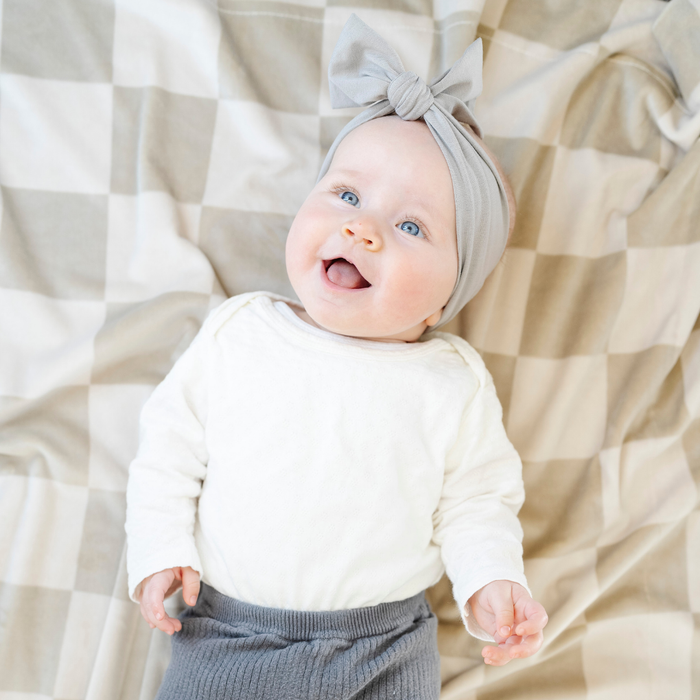 A happy baby girl lays on a neutral checkered minky stretch blanket. The soft blanket is a luxury blanket and a Saranoni blanket. This cozy blanket is also a receiving blanket and is a cute and cozy baby blanket. This stretchy blanket is a great nursery item.