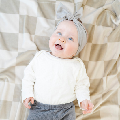 A happy baby girl lays on a neutral checkered minky stretch blanket. The soft blanket is a luxury blanket and a Saranoni blanket. This cozy blanket is also a receiving blanket and is a cute and cozy baby blanket. This stretchy blanket is a great nursery item.