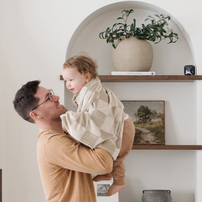 A father holds his little boy who is wrapped in a neutral checkered minky stretch blanket. The soft blanket is a luxury blanket and a Saranoni blanket. This cozy blanket is also a receiving blanket and is a cute and cozy baby blanket. This stretchy blanket is a great nursery item.