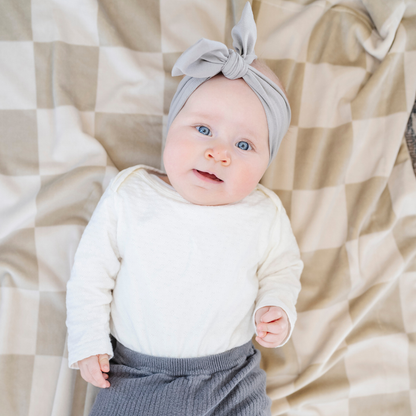 A baby girl lays on a neutral checkered minky stretch blanket. The soft blanket is a luxury blanket and a Saranoni blanket. This cozy blanket is also a receiving blanket and is a cute and cozy baby blanket. This stretchy blanket is a great nursery item.