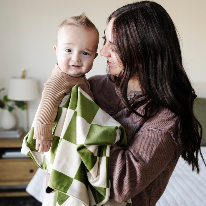 A mother holds her happy baby boy and a olive and cream checkered minky stretch blanket. The soft blanket is a luxury blanket and a Saranoni blanket. This cozy blanket is also a receiving blanket and is a cute and cozy baby blanket. This stretchy blanket is a great nursery item.