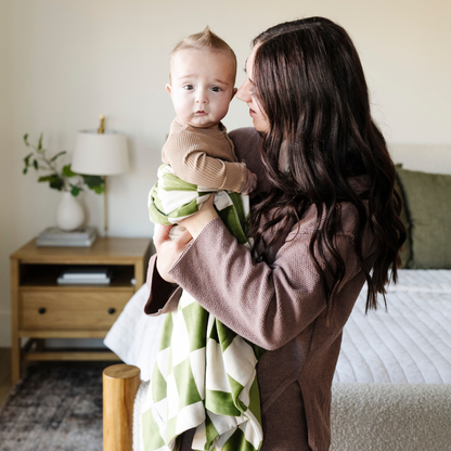 A mother holds her baby boy and a olive and cream checkered minky stretch blanket. The soft blanket is a luxury blanket and a Saranoni blanket. This cozy blanket is also a receiving blanket and is a cute and cozy baby blanket. This stretchy blanket is a great nursery item.