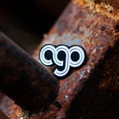 Enamel pin that has the monogram AGO spelled out in white font with a grey line through the letters. The pin is sitting on a rusty piece of metal.