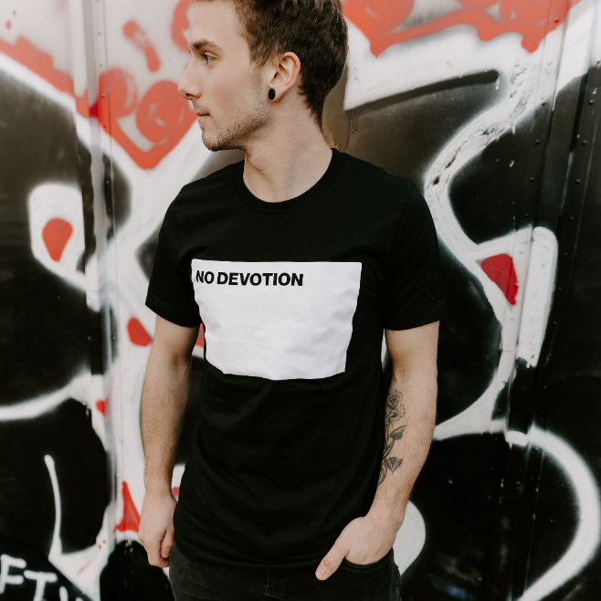 A young man with short hair and a tattoo on his left arm stands before a black and red graffiti wall, wearing a No Devotion black T-shirt featuring "NO DEVOTION" inside a white rectangle.