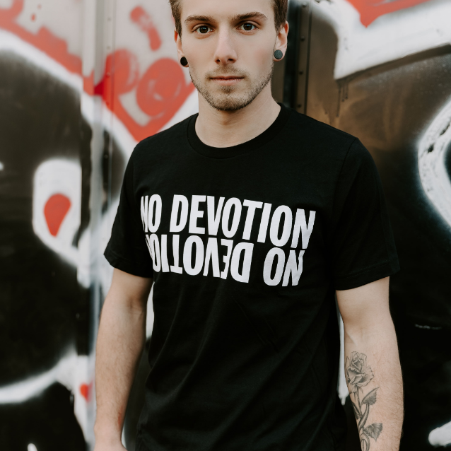 A young person with short hair and a tattoo stands before a graffiti-covered wall, wearing a No Devotion black T-shirt with NO DEVOTION boldly printed upside down in white.