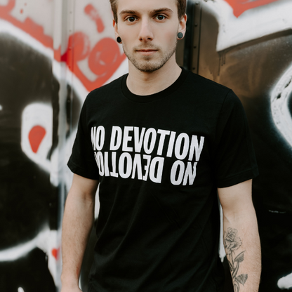 A young person with short hair and a tattoo stands before a graffiti-covered wall, wearing a No Devotion black T-shirt with NO DEVOTION boldly printed upside down in white.