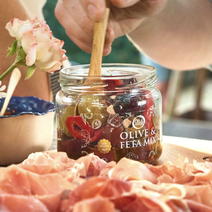 A lifestyle shot of a jar of Olive & Feta Mix, showing green and black olives along with pieces of feta cheese, marinated in oil. Cured meats sit on a wooden board in front of the jar.