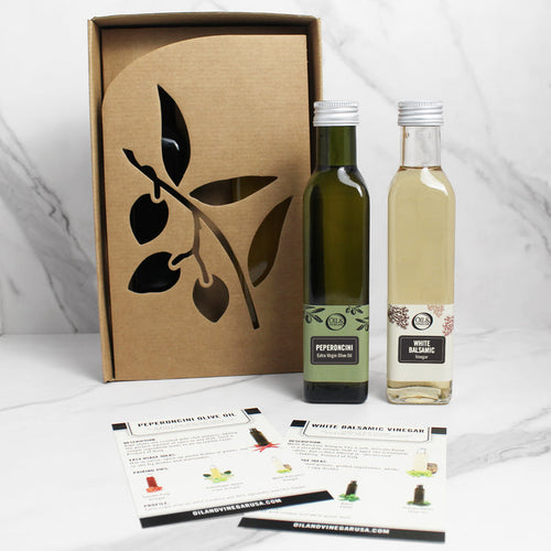 A bottle olive oil and white balsamic vinegar placed outside a cardboard box with a leaf design. The vinegar bottle is labeled with 'White Balsamic' and the olive oil bottle with 'Peperoncini Extra Virgin'.