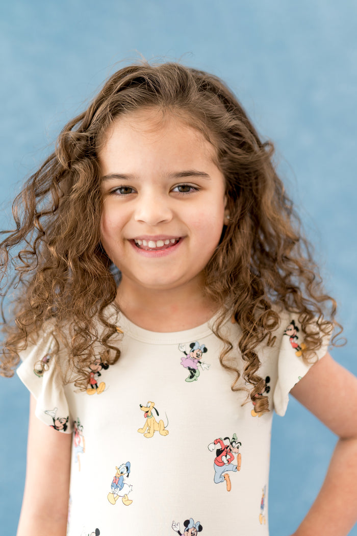 Young girl with curly hair wearing a cream dress with cartoon character prints against a blue background