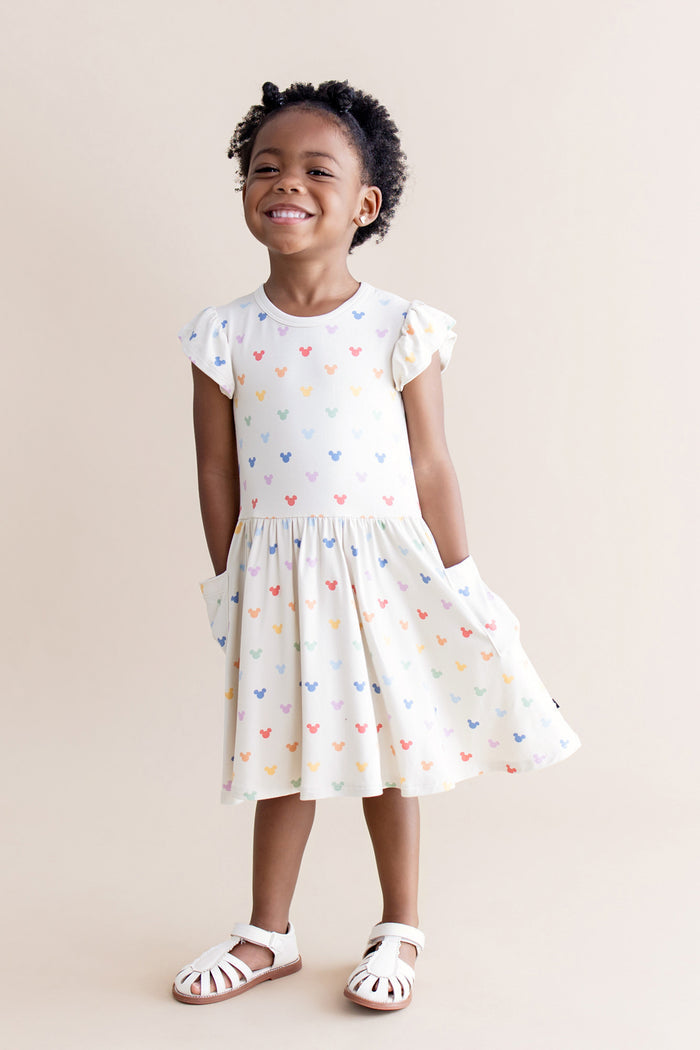 Young girl wearing a cream pocket dress with colorful mickey patterns on a beige background