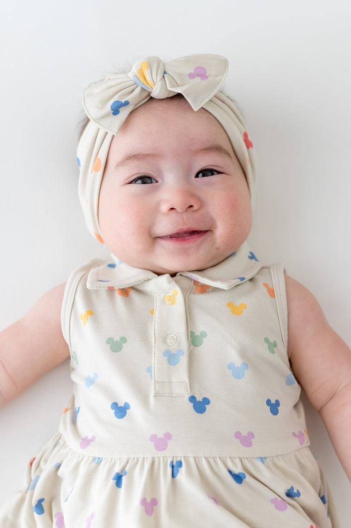 Baby wearing a patterned dress and headband with colorful bows on a plain background