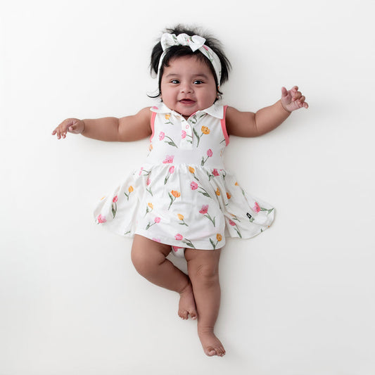 Young infant laying on a white surface wearing the Polo Bodysuit Dress in Tulip with matching bow headband