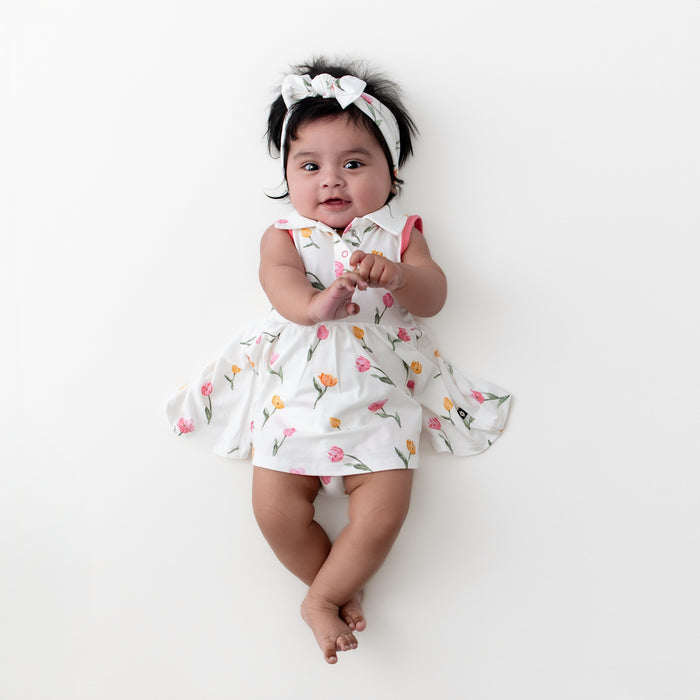 Infant laying on a white surface wearing the Polo Bodysuit Dress in Tulip with matching knotted bow headband