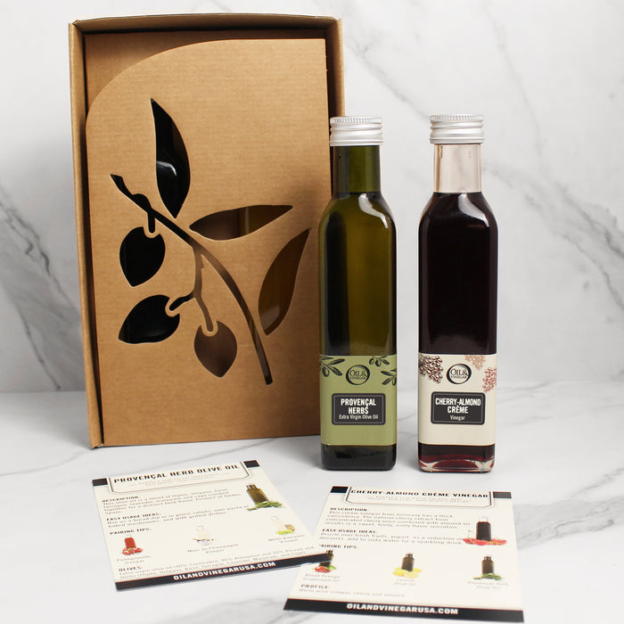 Two bottles of vinegar and a bottle of olive oil, placed next to a cardboard box with a cut-out design of leaves. The vinegars are labeled as 'Cherry-Almond Crème' and the olive oil as 'Provencal Herb'.