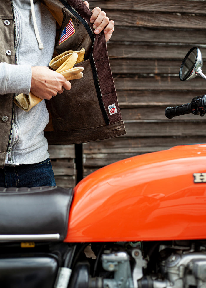 Man in a jacket holding gloves beside an orange Honda motorcycle, with a wooden wall in the background.
