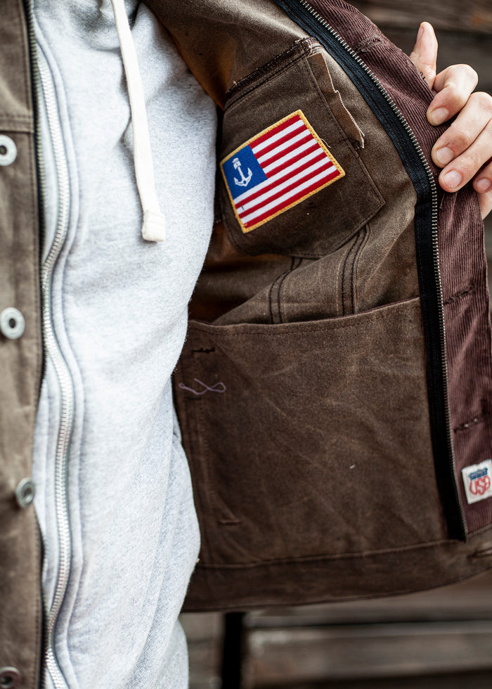 Jacket interior with a pocket holding a patch featuring a flag and anchor. Person wears a gray hoodie, holding jacket open.