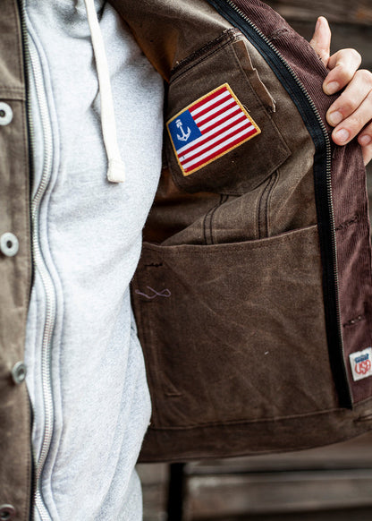 Jacket interior with a pocket holding a patch featuring a flag and anchor. Person wears a gray hoodie, holding jacket open.