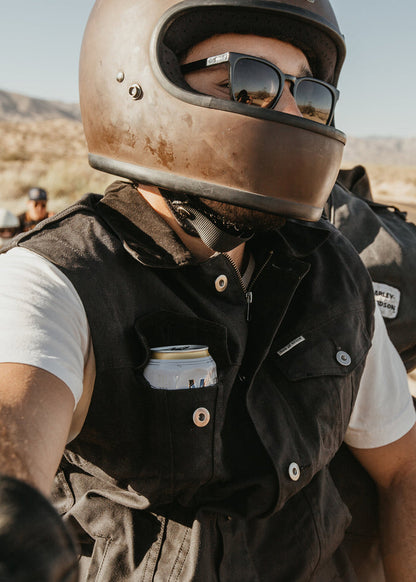 Motorcyclist wearing a helmet and sunglasses, with a can in his vest pocket, rides through a desert landscape under clear skies.