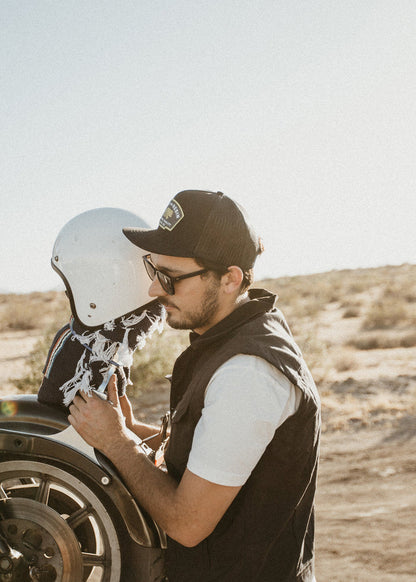 Man in cap and sunglasses adjusts a white helmet on a motorcycle in a sunny desert landscape, creating a serene and adventurous vibe.