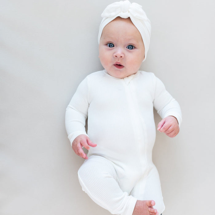 Baby laying on eggshell colored surface wearing Kyte Baby Ribbed turban style Headwrap in Cloud