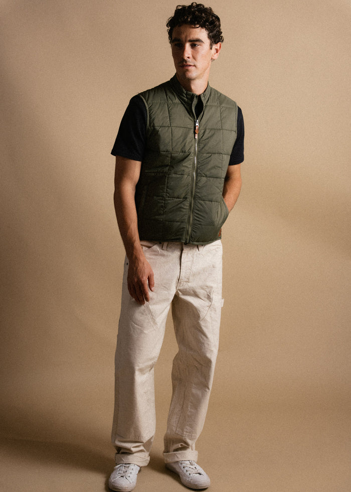Man in olive vest, black shirt, beige pants, and white sneakers stands against a beige backdrop, hands in pockets, looking ahead.
