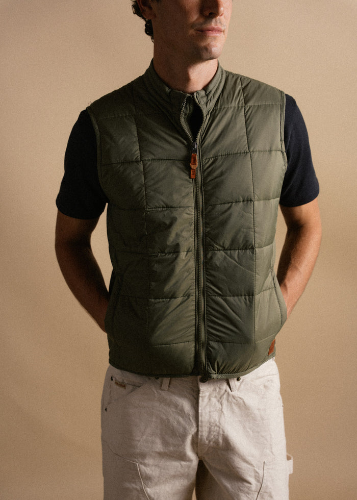 Man in olive quilted vest and beige pants stands against a neutral background, hands in pockets, exuding casual style.