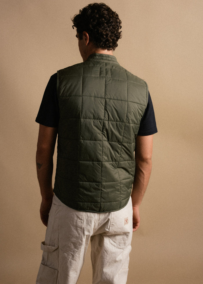 Man in green quilted vest and white cargo pants stands against a beige background, facing away, showcasing casual style.