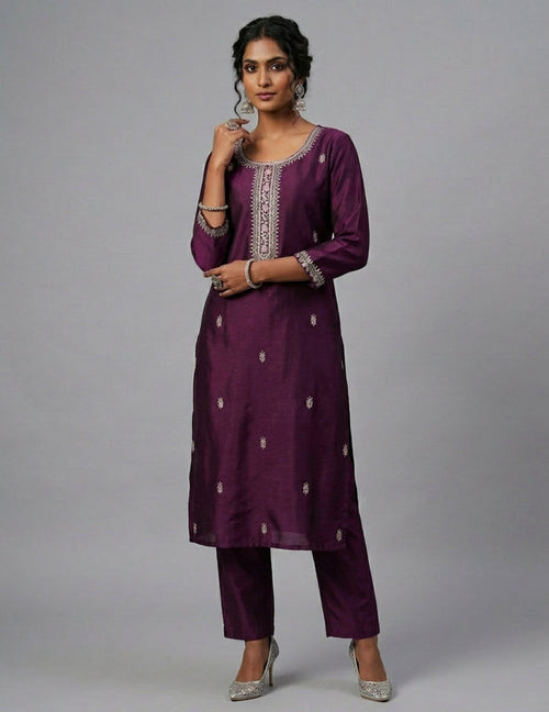 A woman modeling a deep purple Indian ethnic outfit consisting of a long kurta with silver embroidery and matching trousers, paired with silver high heels, against a solid gray studio background.