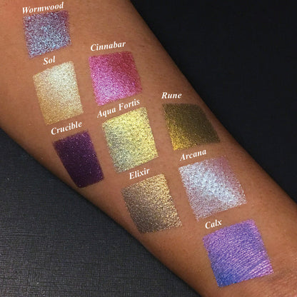 Top angled arm swatches on medium skin tone of Rune Duochrome Eyeshadow next to Wornwood, Sol, Cinnabar, Crucible, Aqua Fortis, Elixir, Arcana, Calx